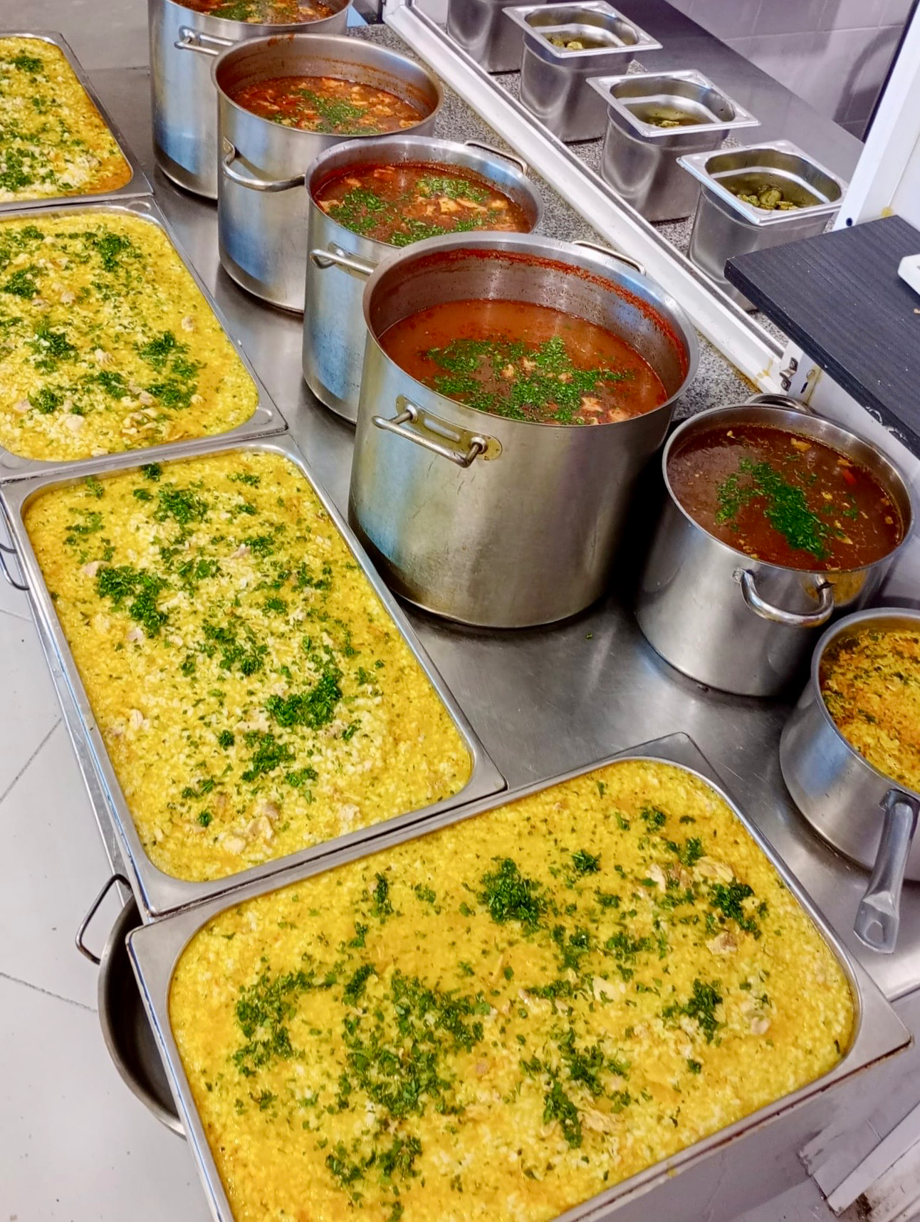 Tăvi mari cu pilaf cu carne și ierburi verzi alături de oale din inox cu supă roșie de legume, pregătite în bucătăria centrului de îngrijire pentru seniori