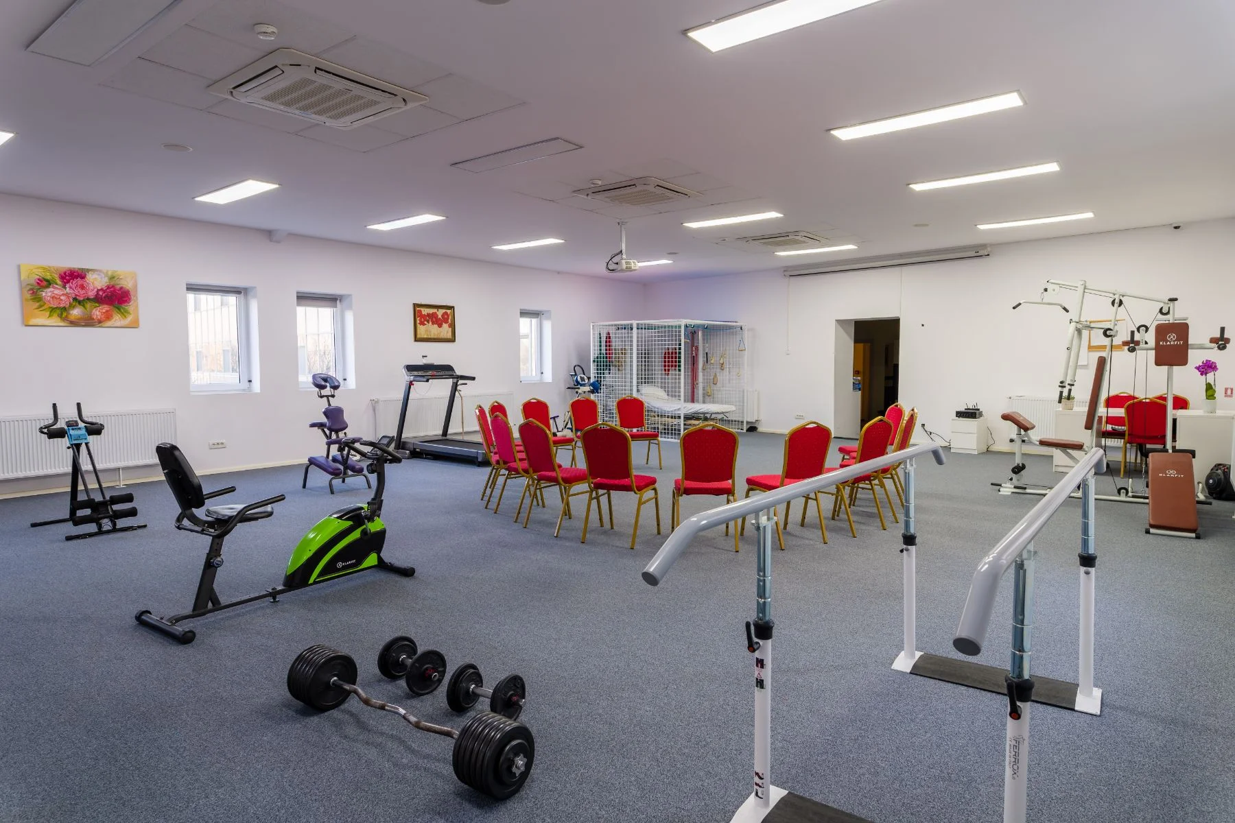 Sala de kinetoterapie cu aparate de fitness, scaune rosii, gantere si echipamente medicale pentru reabilitare seniori
