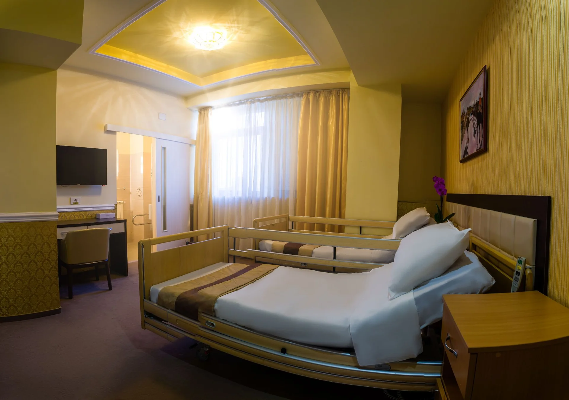 Camera moderna pentru seniori cu pat medical reglabil, TV pe perete, mobilier elegant si ferestre mari cu perdele galbene