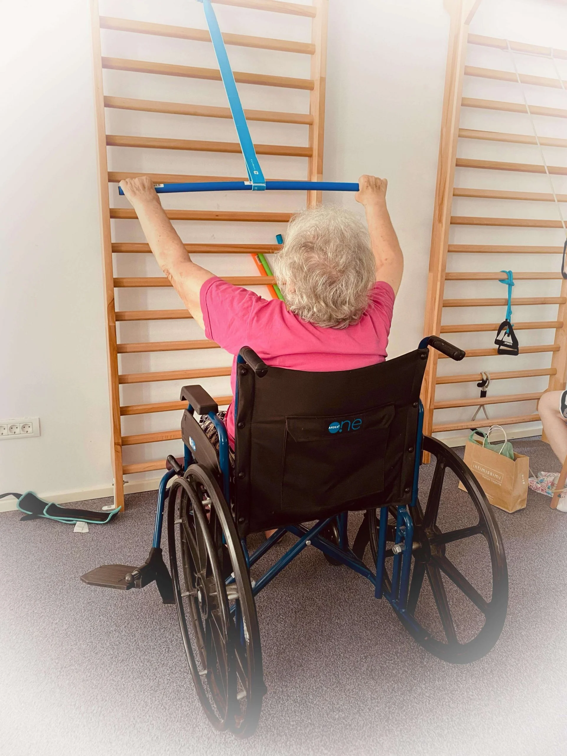 Senior în scaun cu rotile efectuând exerciții de kinetoterapie cu o bandă elastică albastră la barele de gimnastică