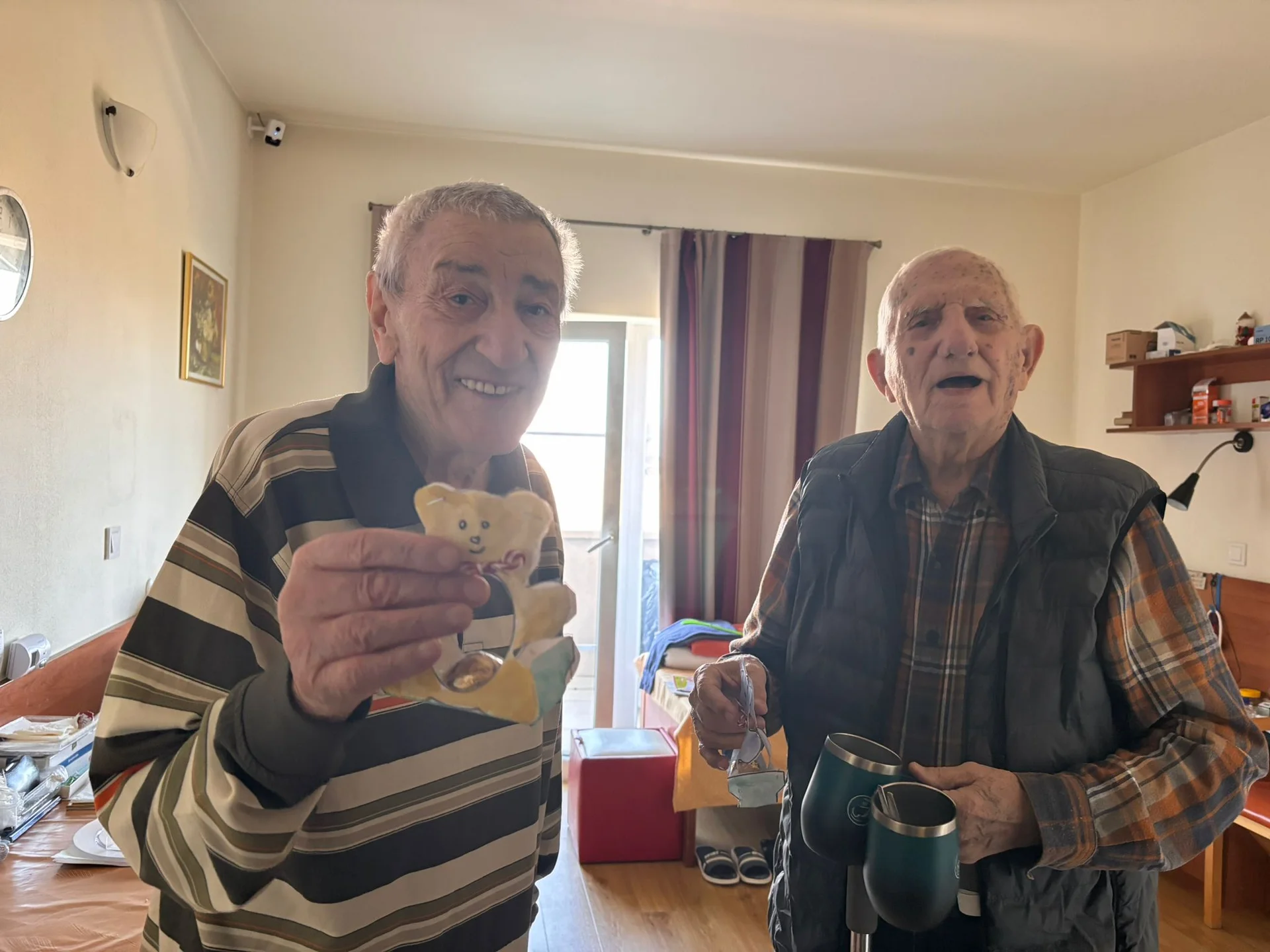 Doi bărbați în vârstă zâmbitori într-o cameră, unul ținând o jucărie de ursuleț, celălalt o cană, participând la activități recreative