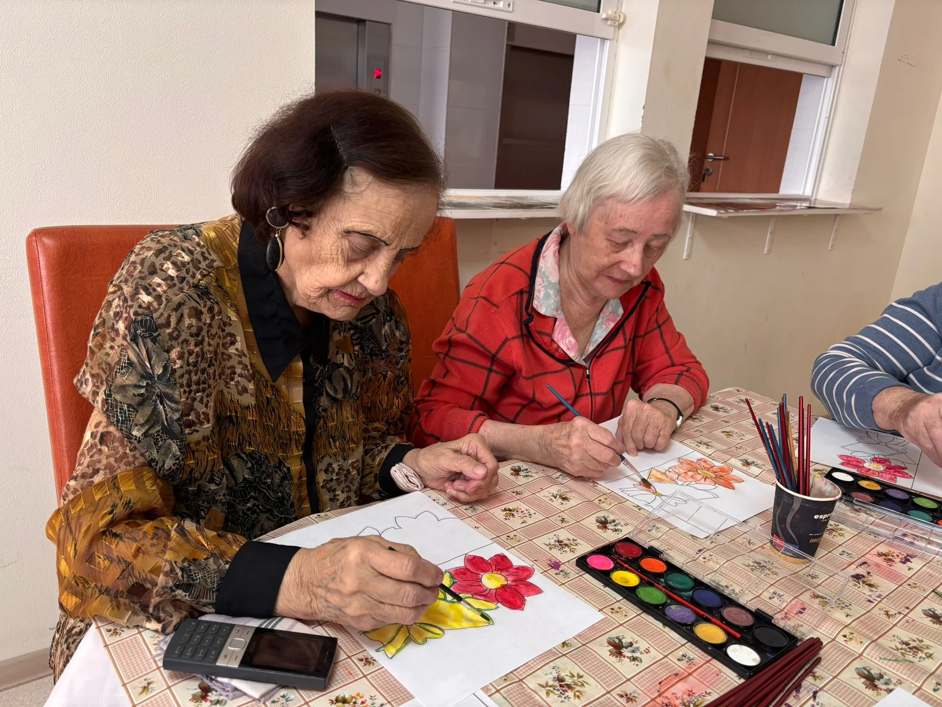 Două femei în vârstă pictând flori cu acuarele la o masă într-un centru de îngrijire seniori