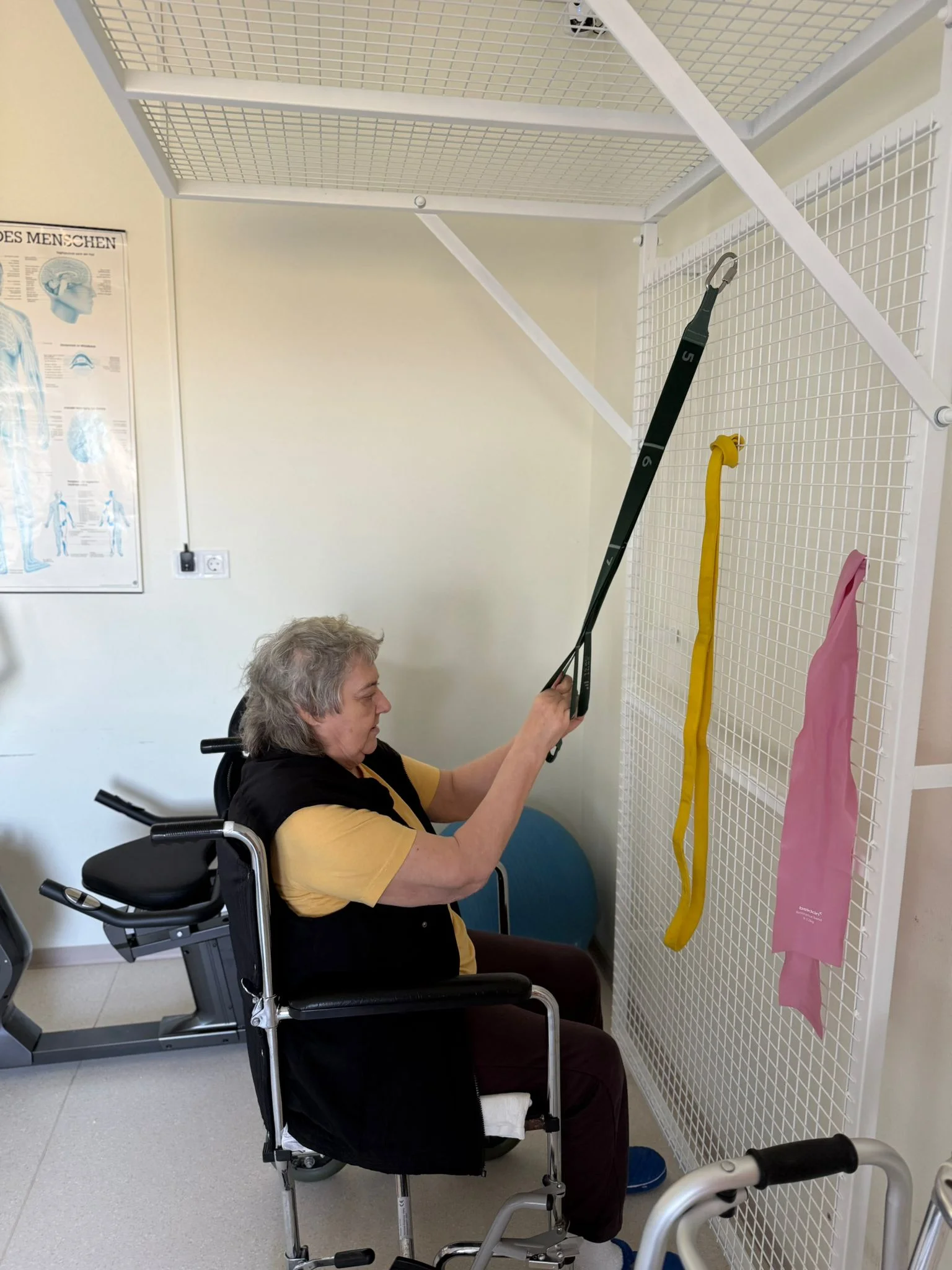 Senior in scaun cu rotile efectuand exercitii de kinetoterapie cu benzi elastice la un aparat de recuperare