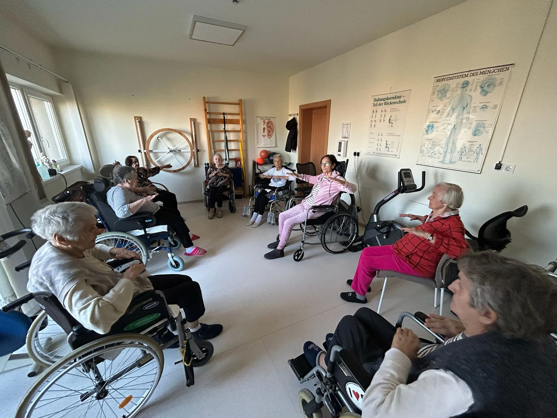 Seniori in scaune rulante participa la o sedinta de kinetoterapie intr-o sala cu echipamente de recuperare si diagrame medicale pe pereti
